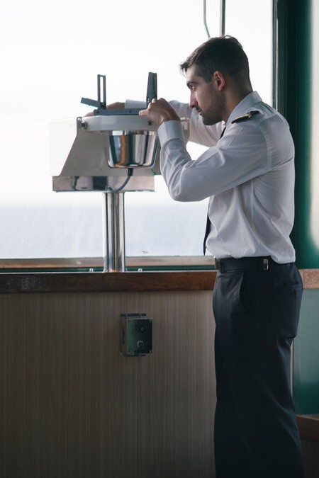 A uniformed ship officer stands indoors, looking through a navigational instrument mounted by a window, possibly using a sextant or alidade for observations.