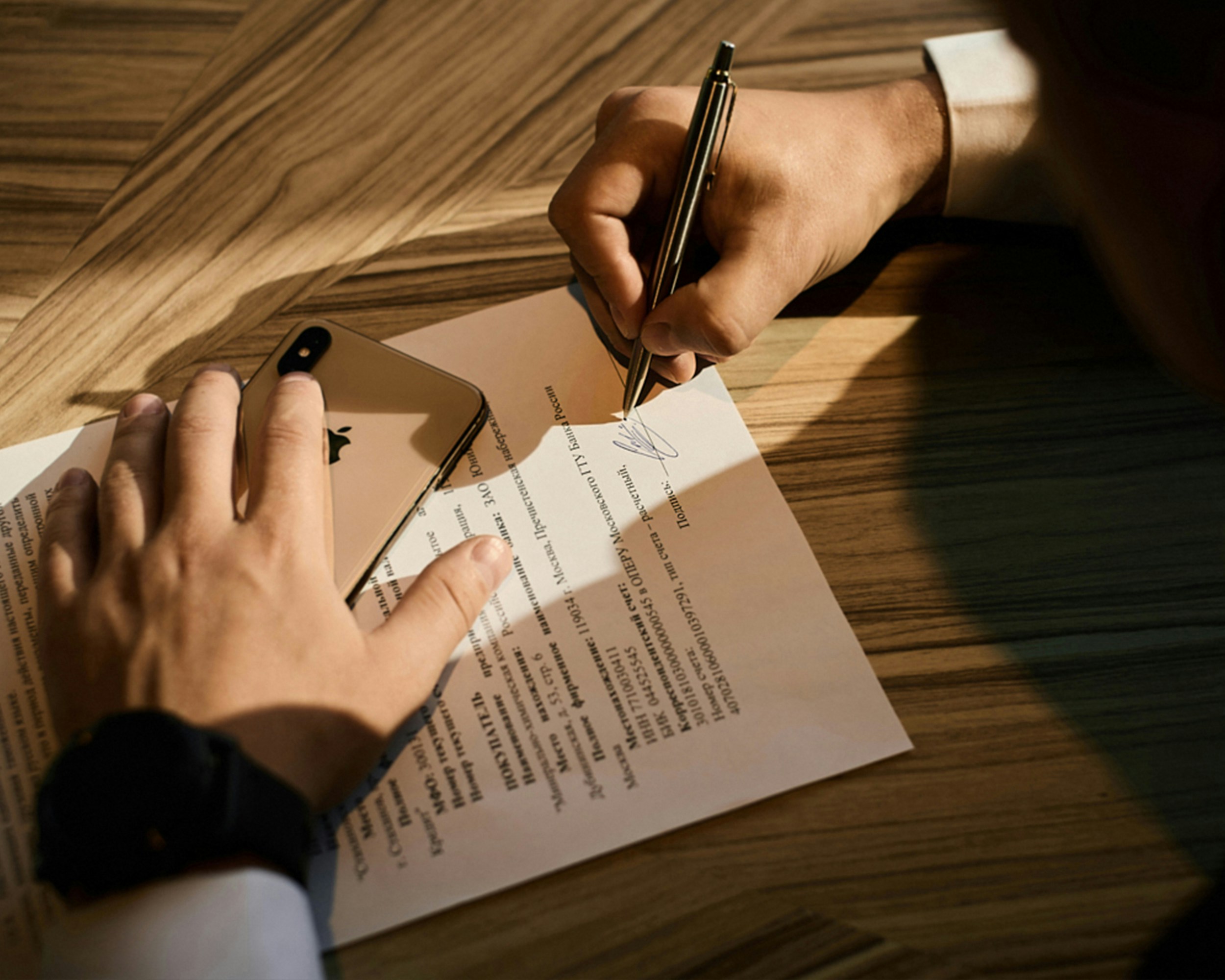 A person signs a document with a pen on a wooden table. Their other hand holds a mobile phone beside the paper. The scene is well-lit and focused on the hands and paper.