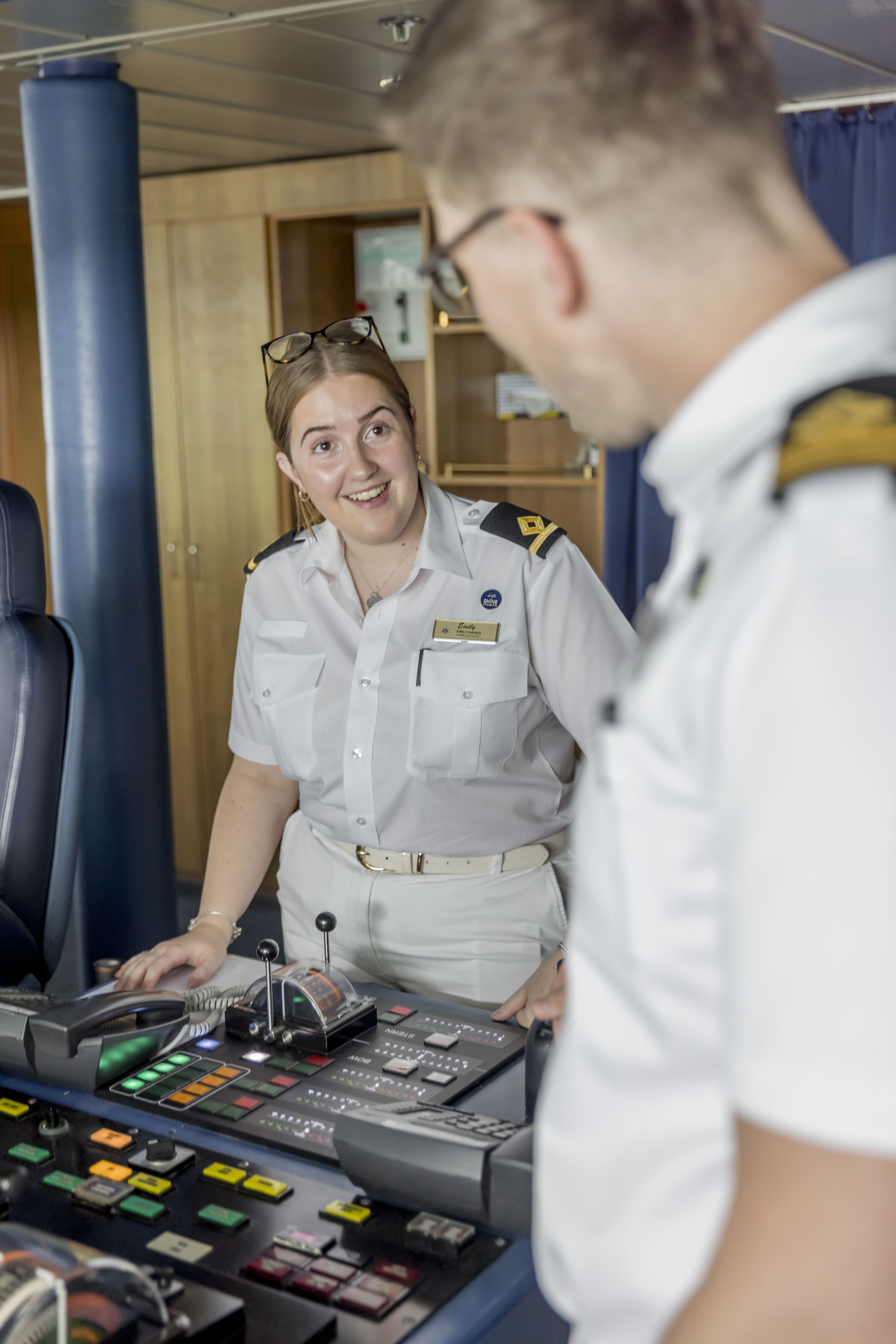 A woman in a white uniform, smiling and talking to a male colleague, stands at a control panel with various buttons and levers inside a ship’s bridge.