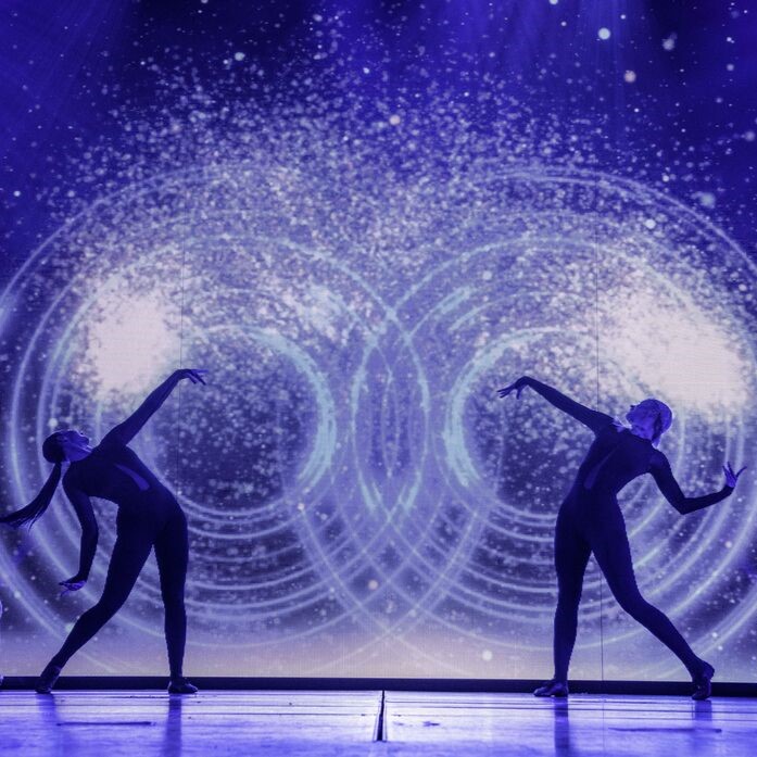 Two dancers in dark bodysuits pose dramatically on stage, leaning back with arms extended, against a backdrop of swirling, glowing patterns and bright particles resembling cosmic or galactic imagery.