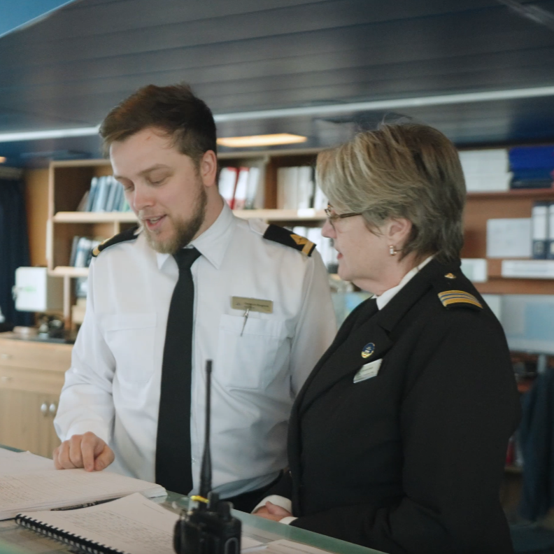 Two people in uniform stand at a desk, reviewing documents together in an office or control room with shelves of books and folders—showing the teamwork essential for P&O Cruises deck careers. A radio sits on the desk in front of them.