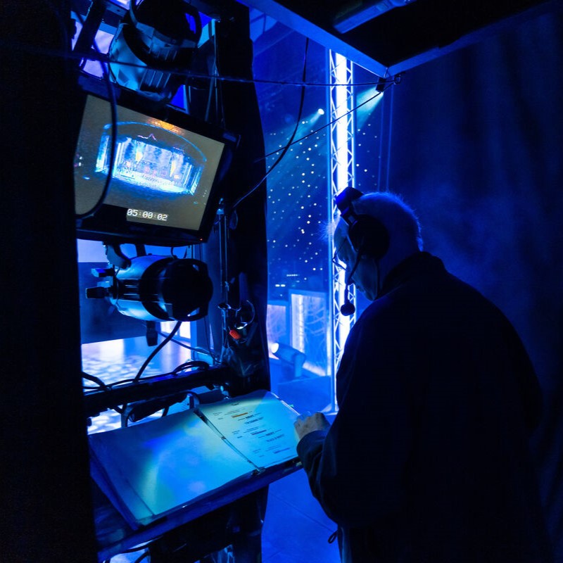 A person with headphones stands backstage next to a monitor and lighting equipment, reviewing a script under blue stage lights, preparing for a live performance or broadcast.