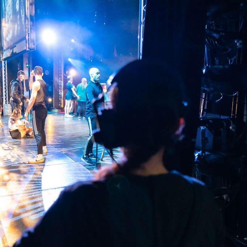 A person wearing a headset stands backstage, watching a group of performers in athletic wear practising or preparing under bright stage lights. The atmosphere is busy and energetic.