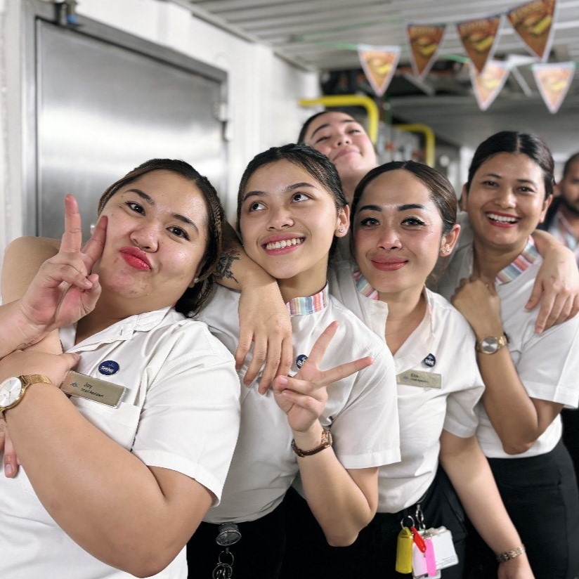 Five women in white uniforms smile and pose together for the camera, making peace signs and standing closely in what appears to be a workplace or staff room with decorations in the background.