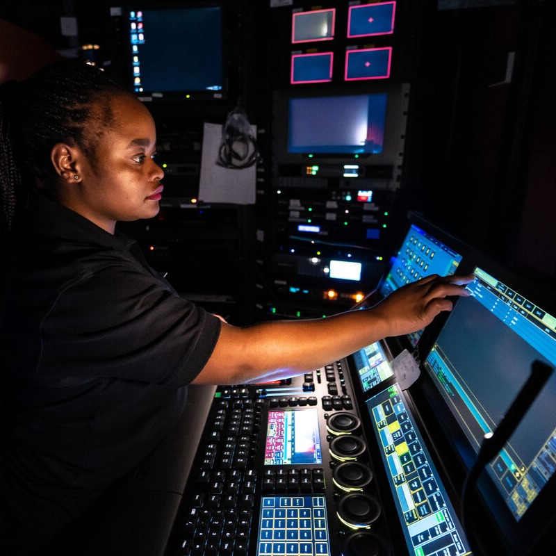 A person operates a complex control panel with multiple monitors and illuminated buttons in a dark room, focusing intently whilst pressing a button on one of the touchscreens.