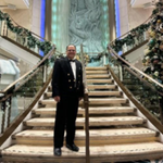 A man in a black dinner jacket stands on a grand, ornately decorated staircase with elegant railings and festive garlands, inside a luxurious building with a large sculpted wall in the background.