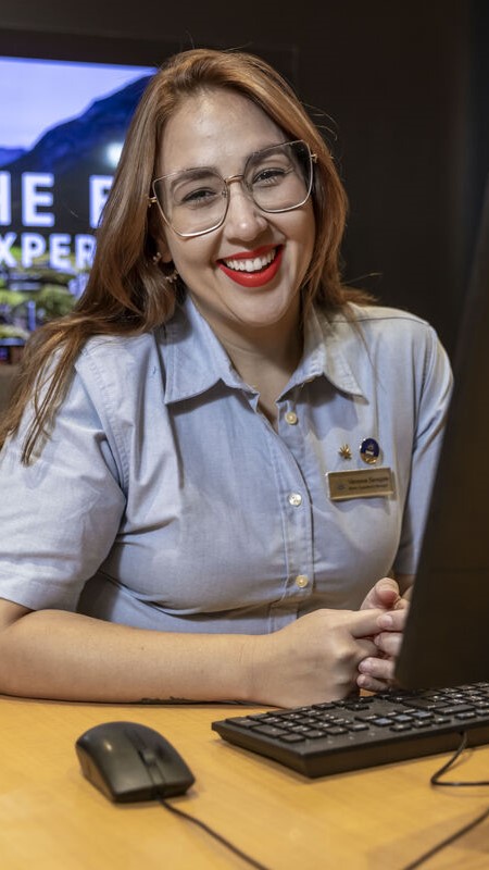A woman with long brown hair, glasses, and red lipstick smiles whilst sitting at a desk with a computer keyboard and mouse. She wears a light blue shirt and a name badge. A screen is visible in the background.