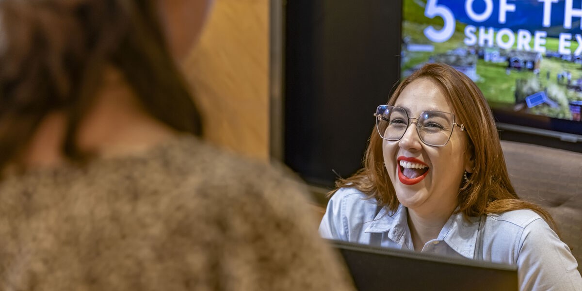 A woman wearing glasses and a light blue shirt sits at a desk, smiling and laughing whilst talking to another person whose back is to the camera. A colourful screen is visible in the background.