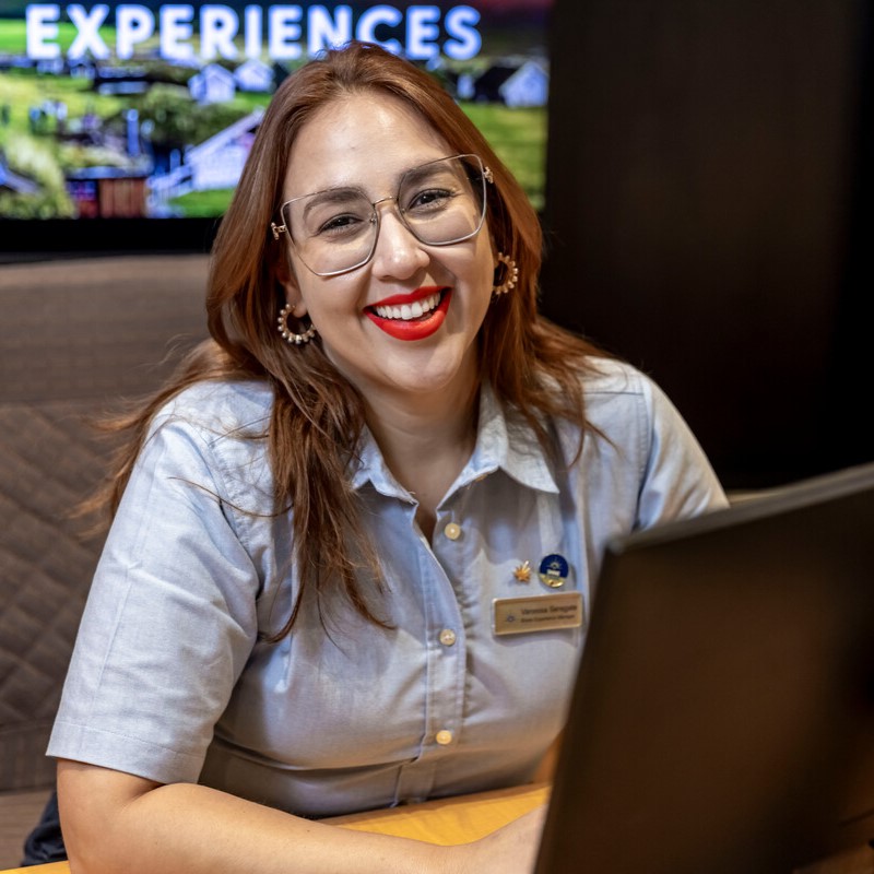 A woman with long brown hair, glasses, and red lipstick smiles while sitting at a desk with a laptop. She wears a light blue blouse and a name badge. A colourful EXPERIENCES sign is visible in the background.