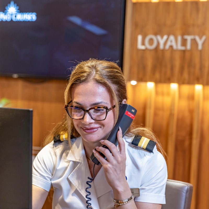 A woman in a white uniform with shoulder epaulettes and glasses is smiling whilst speaking on the phone at a desk. A sign behind her reads LOYALTY on a wooden wall.