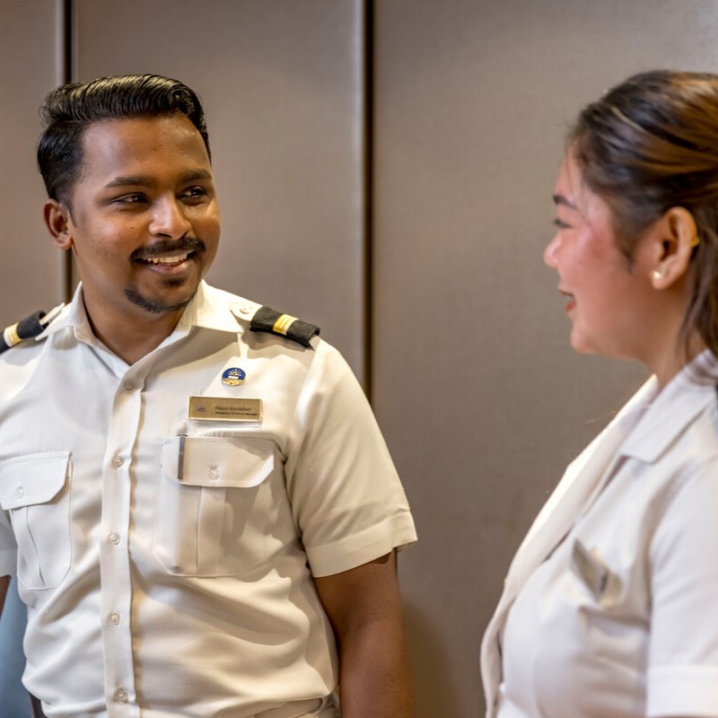 Two people in white uniforms with shoulder epaulettes stand indoors, smiling and having a conversation. The background is plain, emphasising their friendly interaction.