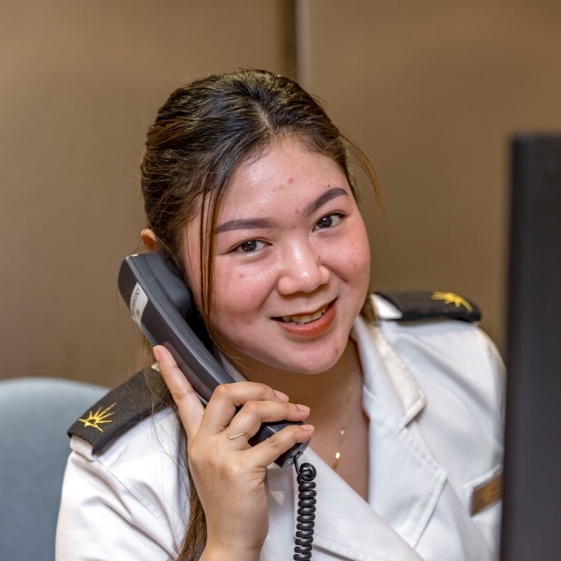 A woman in a white uniform sits at a desk, smiling whilst holding a telephone to her ear. She appears to be engaged in a conversation, looking friendly and professional.