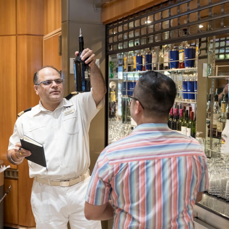 A man in a white uniform holds up a wine bottle behind a bar, speaking to a customer in a striped shirt. Shelves with glasses and bottles are visible in the background.