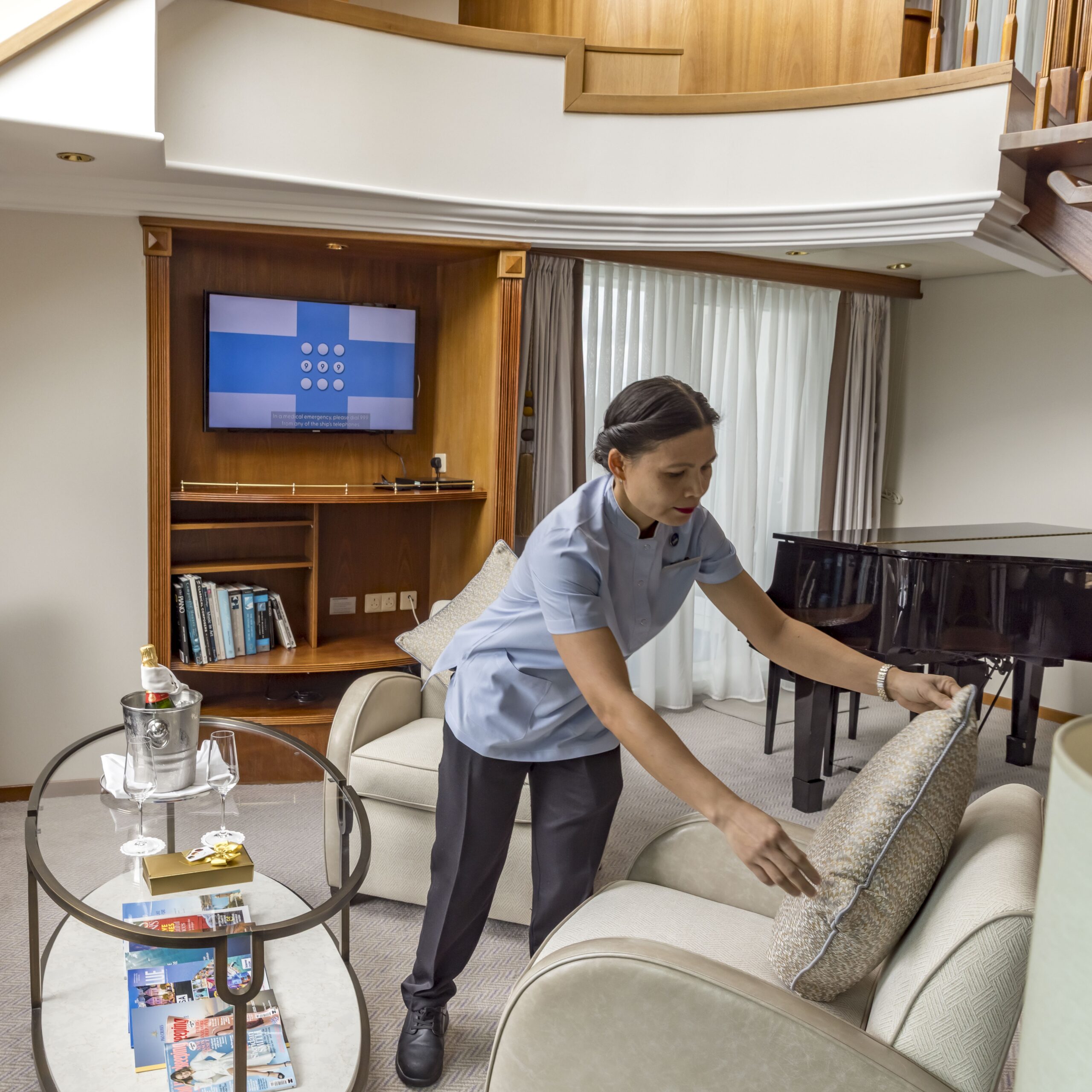 A woman in a housekeeping uniform arranges a cushion on a sofa in a modern, well-lit living room with a TV, bookshelves, a glass coffee table, and a black piano.