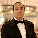 A man dressed in a formal black dinner suit with a bow tie stands indoors in front of a blurred background with warm lighting. He is looking at the camera with a slight smile.