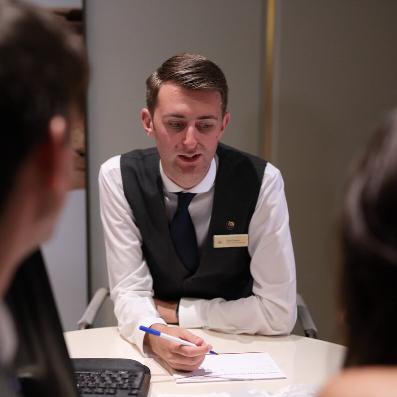 A man in a formal waistcoat and tie sits at a desk, holding a pen and speaking to two people opposite him. Papers and a keyboard are on the table. He appears to be explaining something or giving advice.