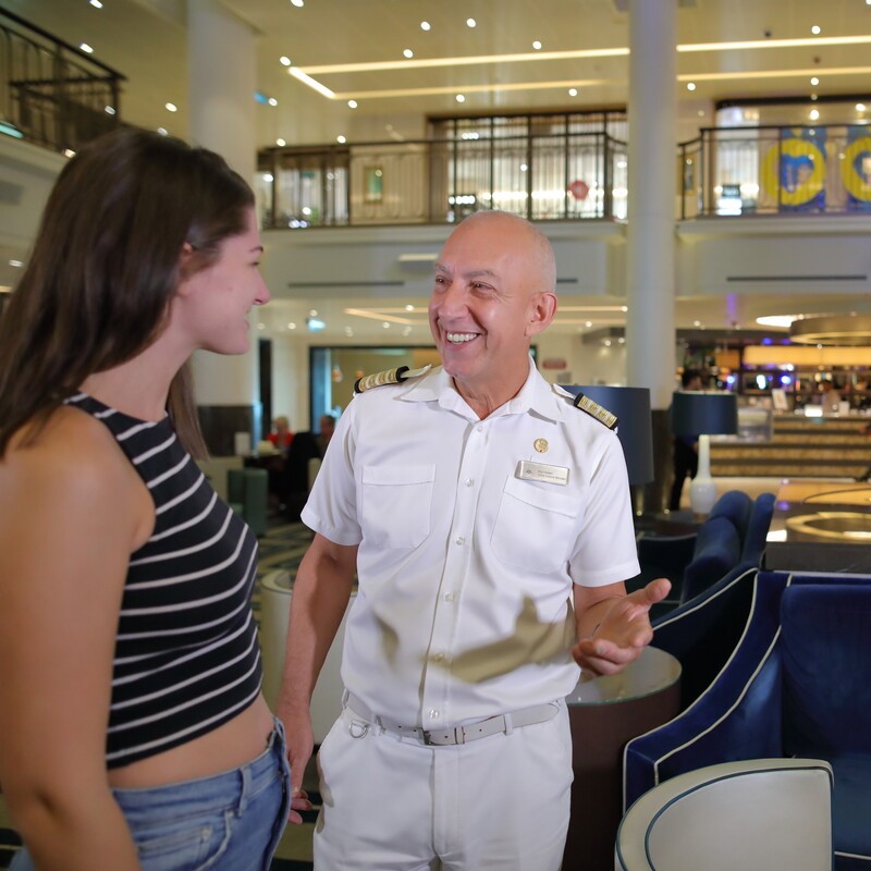 A man in a white captain’s uniform smiles and talks with a young woman in a striped vest top inside a modern, well-lit lobby with blue chairs and a balcony in the background.