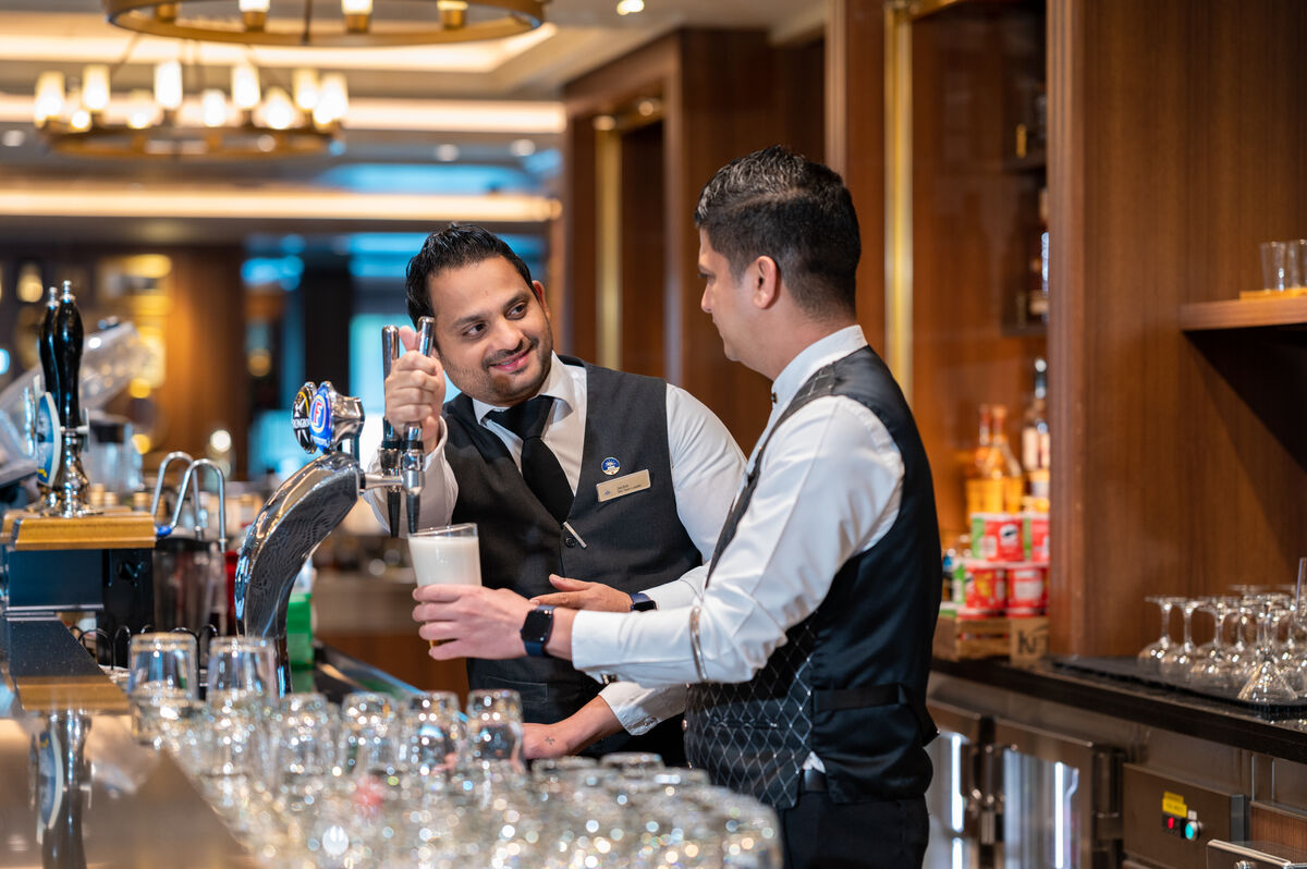 Two bartenders in uniform work behind a bar on a cruise ship; one pours a draught beer into a glass while the other watches and converses with him. Shelves with glassware and drink supplies are visible, typical of cruise ship bar jobs.