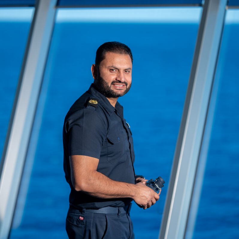 A man in a navy uniform stands by large windows overlooking the sea, holding binoculars and smiling at the camera—a glimpse into P&O Cruises deck careers.