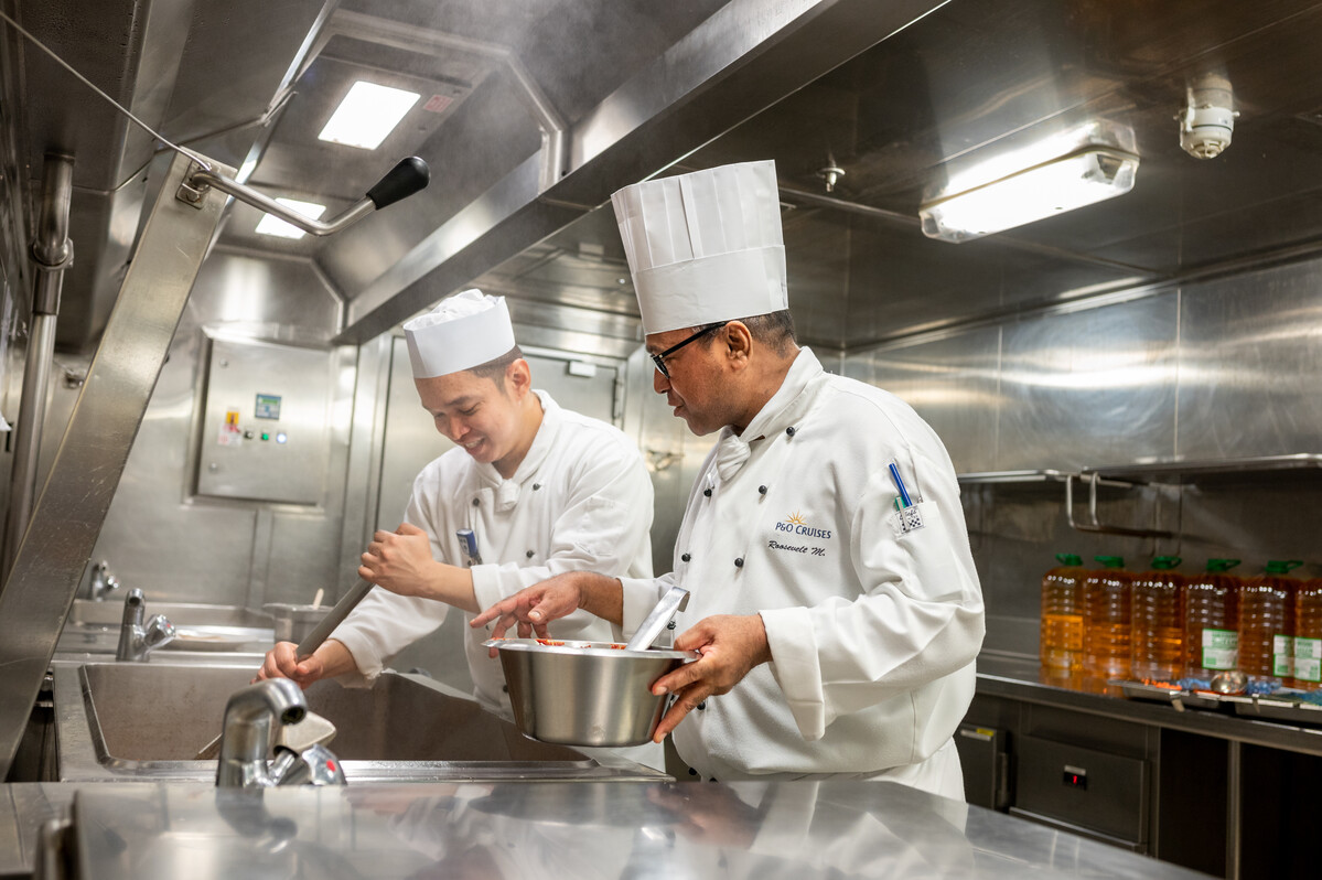 Two chefs in white uniforms and hats work together in a stainless steel commercial kitchen, typical of cruise ship galley jobs, one stirring a bowl and the other washing something at the sink, with bottles of cooking oil lined up in the background.