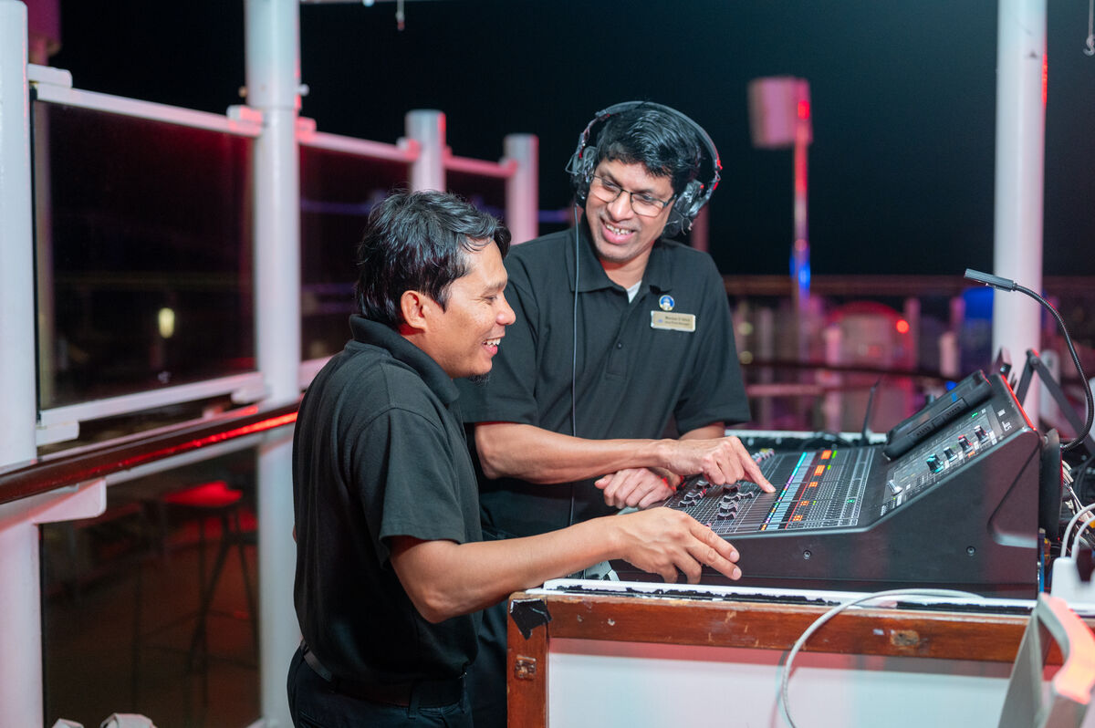 Two men wearing black shirts operate a sound mixer on an outdoor terrace at night. One man, smiling, points at the mixer while the other, wearing headphones and glasses, looks on and smiles.
