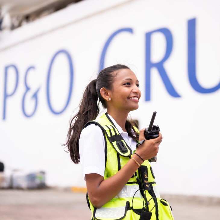 A smiling woman in a reflective vest holds a walkie-talkie, standing outdoors before a white wall with large blue letters that partially read P&O CRU, representing the dynamic environment of P&O Cruises deck careers.
