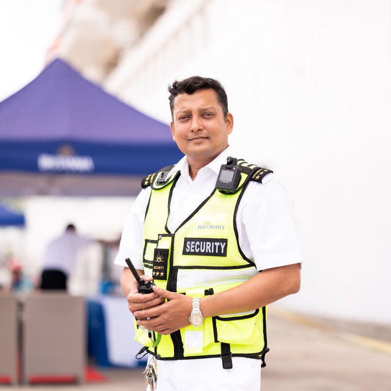 A security guard in a yellow vest and white uniform stands outdoors, holding a walkie-talkie. He could be overseeing P&O Cruises deck careers, with a blue marquee, blurred people, and a white wall in the background.