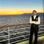A person in a formal waistcoat and tie stands on the deck of a ship at sunset, with the ocean and distant mountains in the background.