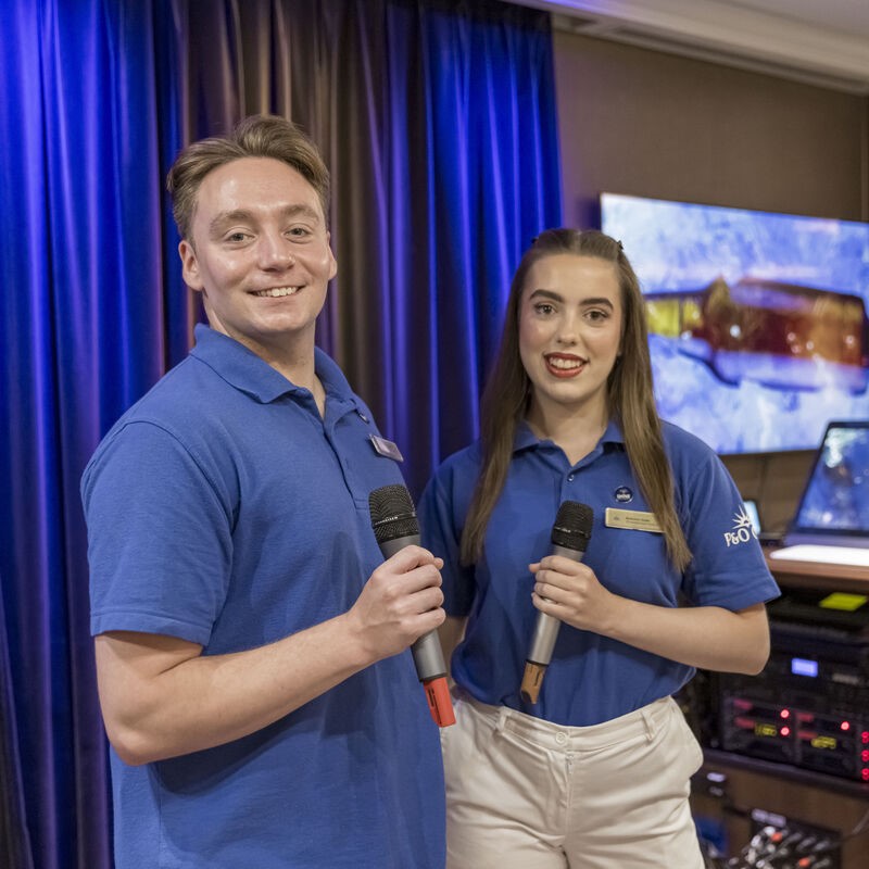Two young adults in blue polo shirts hold microphones and smile at the camera, standing in front of dark blue curtains and audio equipment in a room with a screen displaying a colourful image.