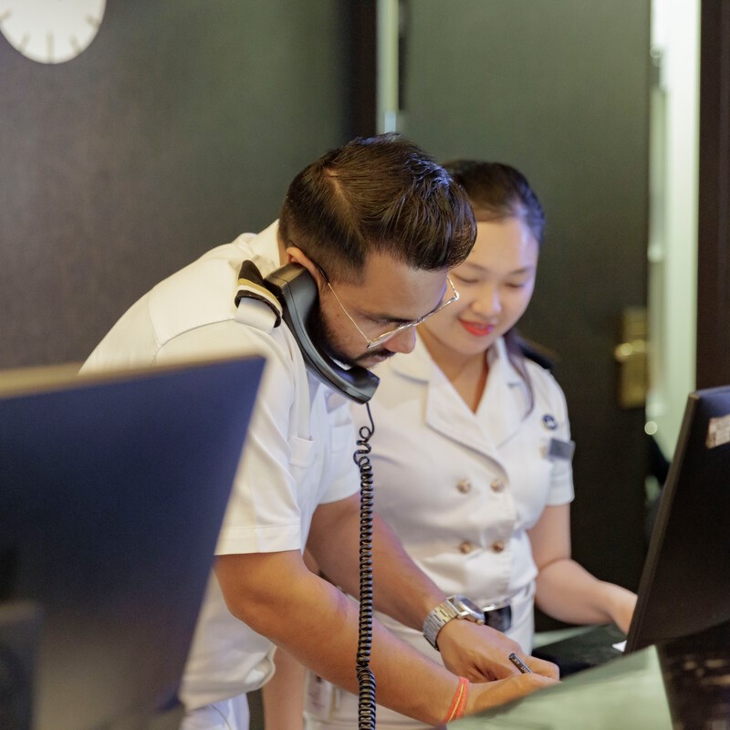 Two people in white uniforms stand at a desk. One man is talking on the phone while writing notes; a woman beside him is looking at a computer screen and smiling. They appear to be working together in a professional setting.