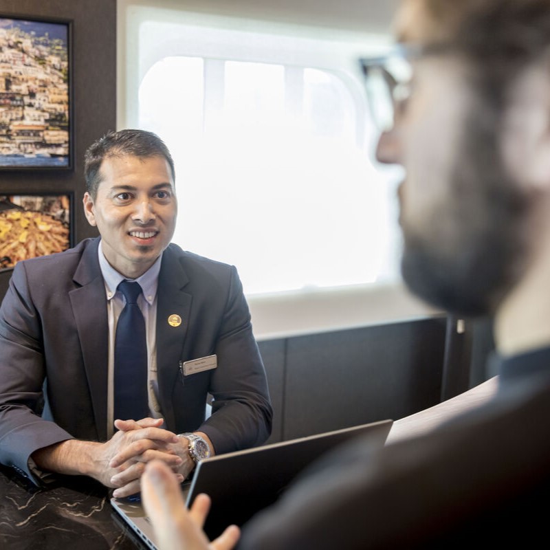 A man in a suit sits at a desk, smiling and talking to another person whose face is blurred in the foreground. They appear to be having a professional conversation in an office setting.
