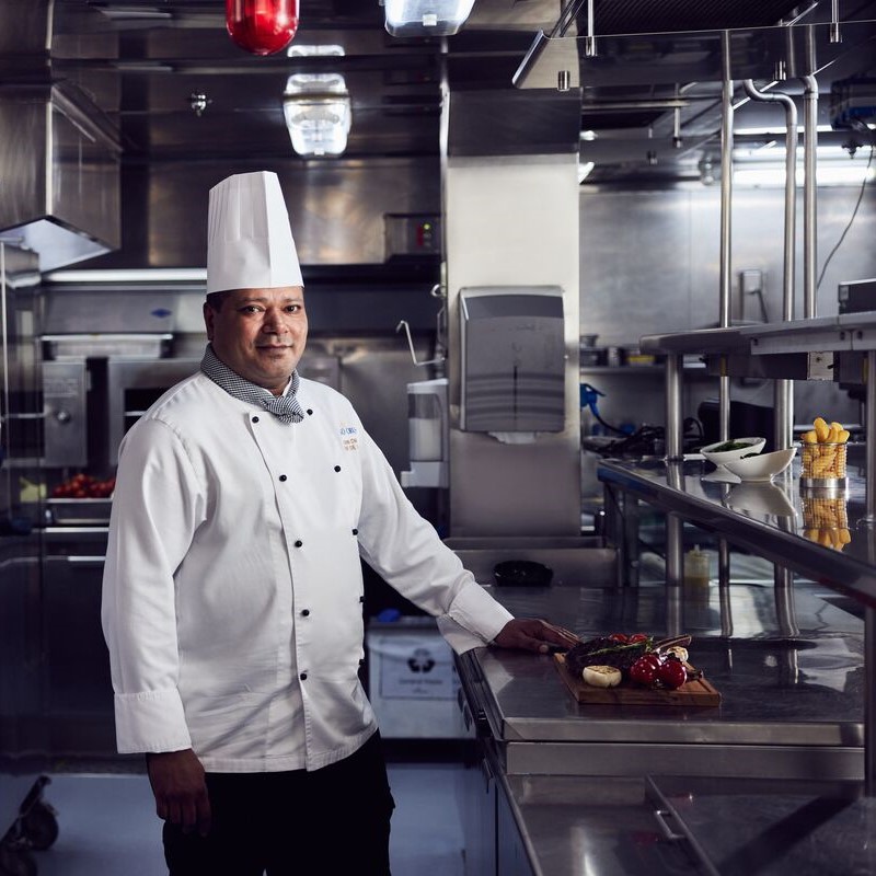A chef in a white uniform and hat stands in a modern, stainless steel cruise ship galley, smiling and resting one hand on a counter with fresh vegetables and herbs arranged on a wooden board, showcasing cruise ship galley jobs.