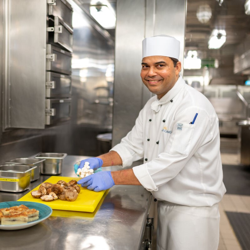 A smiling chef in a white uniform and hat chops mushrooms on a yellow chopping board in a professional kitchen, showcasing the fast-paced environment of cruise ship galley jobs, with various ingredients and prepared food nearby.