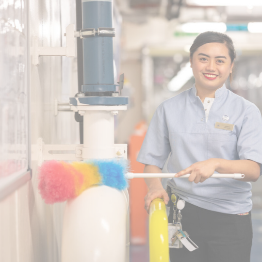 A smiling woman in a light blue uniform dusts a large white pipe with a colourful duster in an indoor setting. She has a name tag and keys attached to her waistband.