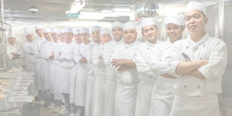 A group of chefs in white uniforms and hats stand side by side in a professional kitchen, reminiscent of cruise ship galley jobs, facing the camera with arms folded, ready for service.