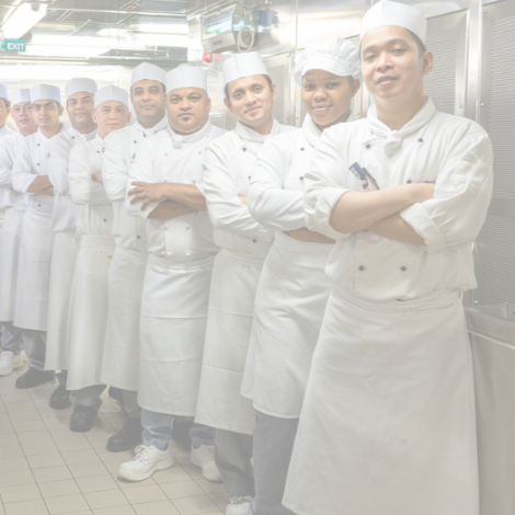 A group of chefs in white uniforms and hats stand in a line in a commercial kitchen, smiling with arms folded. The background features stainless steel equipment, highlighting the teamwork found in cruise ship galley jobs.