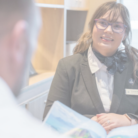 A woman in a suit, wearing glasses and a scarf, smiles whilst talking to a person holding an open magazine or brochure in an office setting.
