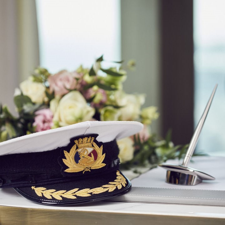A captain’s hat with gold embroidery sits on a table next to a silver pen and a bouquet of white and pink roses, with soft natural light in the background.