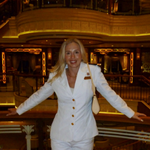 A woman in a white suit stands smiling with arms outstretched on a balcony inside a luxurious, warmly lit building with ornate décor and curved railings.