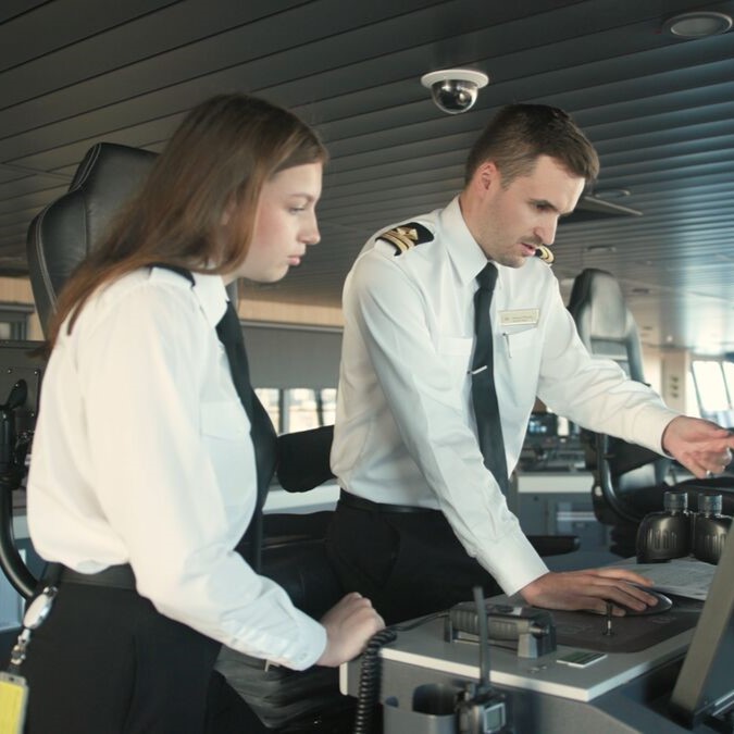 Two ship officers in uniform stand at a control panel on a ship's bridge. One points at a monitor whilst the other observes, both focused on navigation tasks—a glimpse into P&O Cruises deck careers in action.