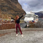 A woman poses with arms outstretched in front of a large cruise ship docked by a fjord, surrounded by mountains and cloudy skies.