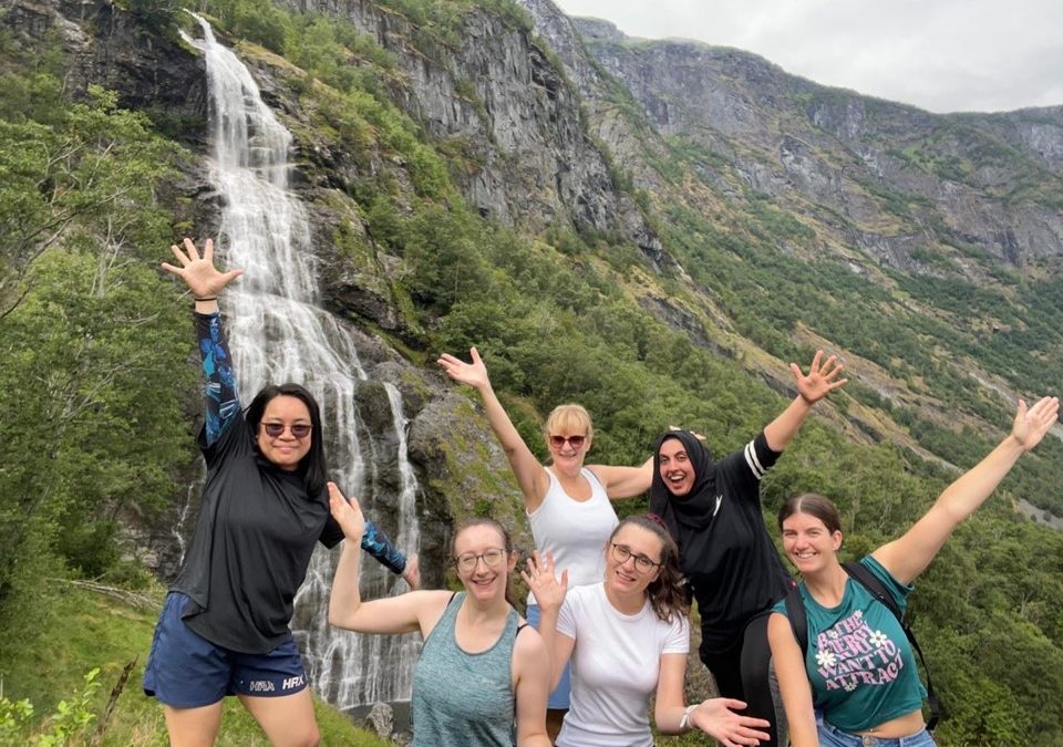 Six smiling women pose with their arms raised in front of a tall waterfall and green, rocky mountains, enjoying an outdoor adventure on a cloudy day.