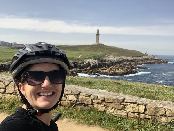 A person wearing sunglasses and a bike helmet smiles at the camera with a rocky coastline and a tall stone lighthouse on a grassy hill in the background.