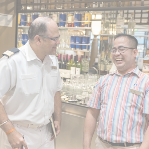 Two men are standing and smiling in front of a bar with shelves of bottles and glasses. One wears a white uniform and holds a notebook, whilst the other wears a striped shirt and spectacles.