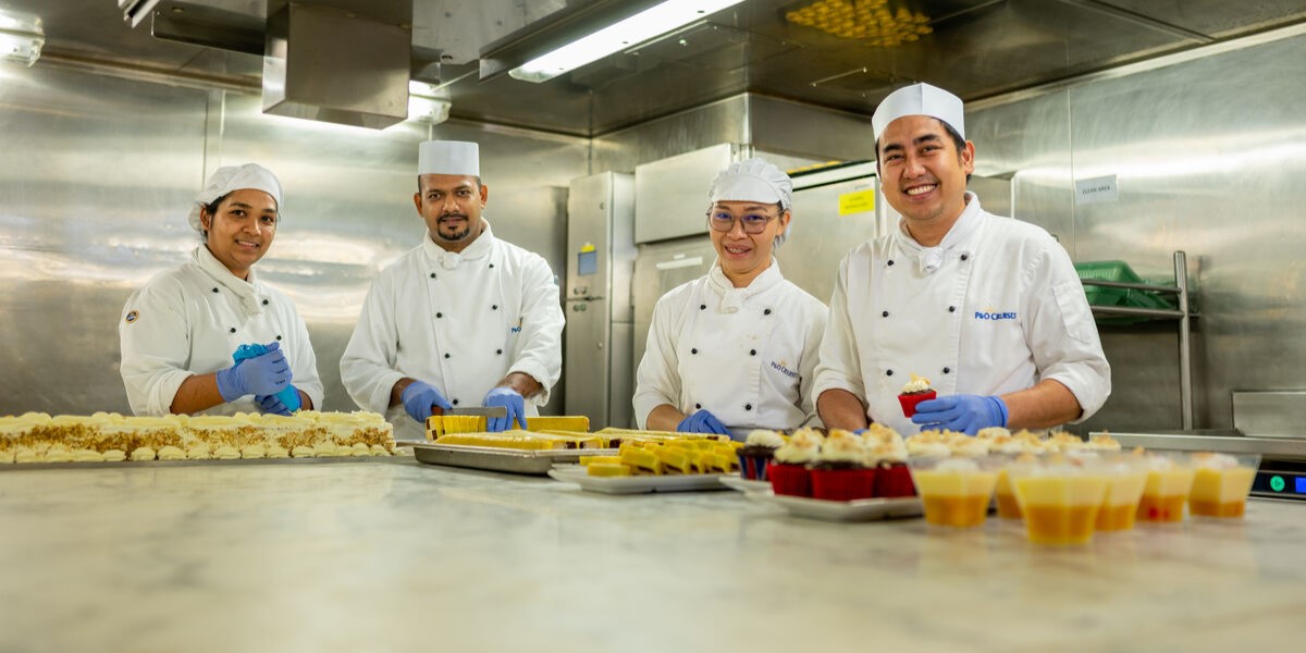Four chefs in white uniforms and hats, wearing blue gloves, stand behind a counter with trays of desserts and pastries in a professional kitchen, smiling at the camera.
