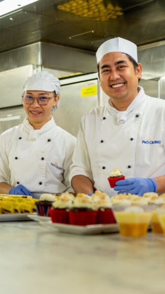 Two smiling chefs in white uniforms and hats, wearing blue gloves, stand in a commercial kitchen with trays of cupcakes and desserts on the counter in front of them.