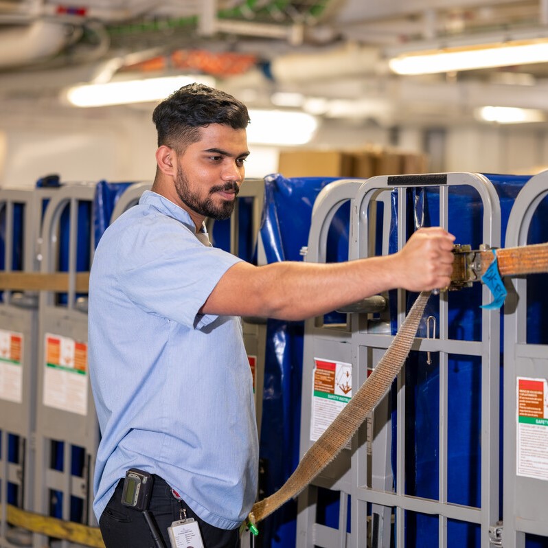 A man in a light blue shirt secures a brown strap around large metal containers in an industrial or warehouse setting.