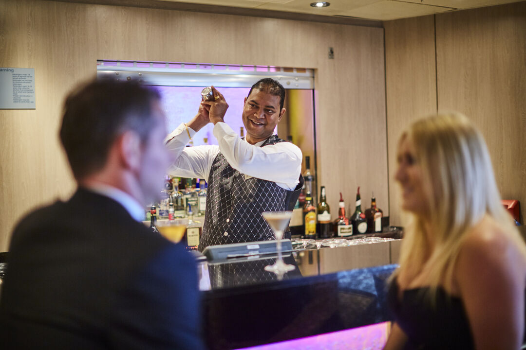 A smiling barman, possibly working one of the sought-after cruise ship bar jobs, shakes a cocktail shaker behind the bar whilst a well-dressed man and woman sit together with drinks. Bottles of alcohol are lined up on shelves behind the barman.