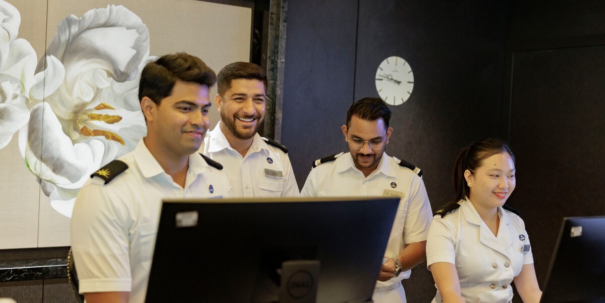 Four uniformed staff members, three men and one woman, stand at computer workstations in a modern office with a flower painting and clock on the wall, smiling and engaged in their work.