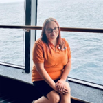A woman with long hair and glasses, wearing an orange shirt and black shorts, sits indoors by a large window overlooking the sea.